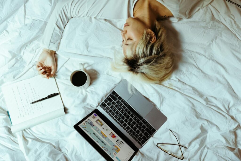 A woman sleeping with a laptop, coffee, and notebook next to her in bed, suggesting a relaxing home environment.