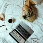 A woman sleeping with a laptop, coffee, and notebook next to her in bed, suggesting a relaxing home environment.