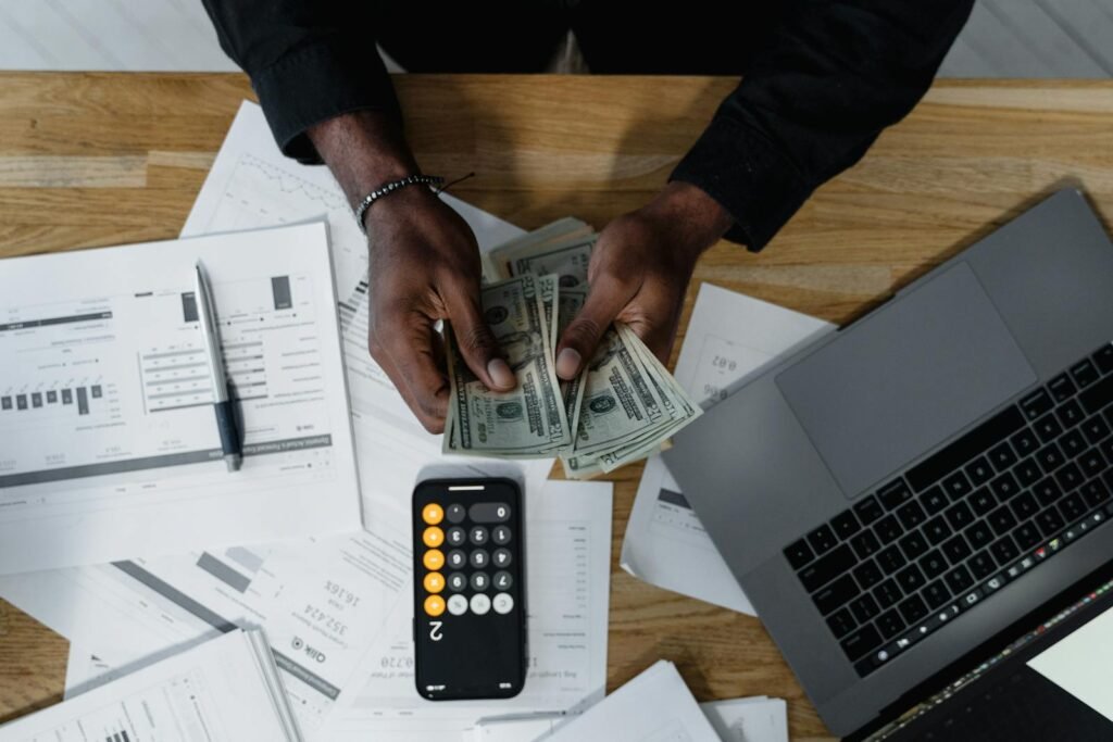 Person counting US dollars, using a calculator and laptop, with financial documents on a wooden desk.