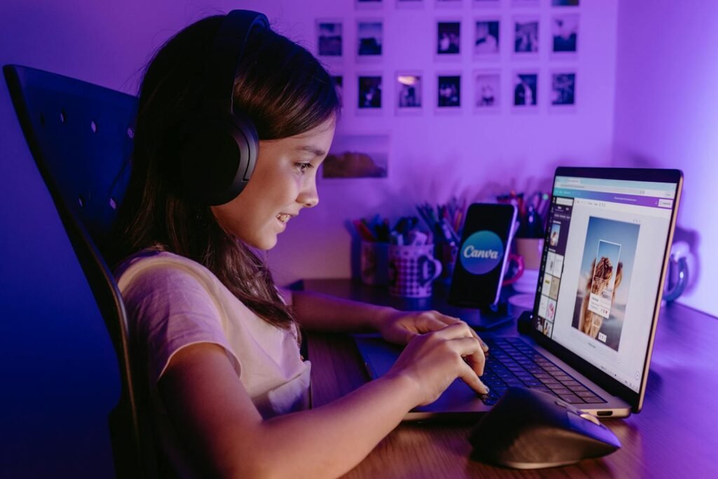 A young girl wearing headphones works on graphic design using a laptop and smartphone in a cozy, creative workspace.